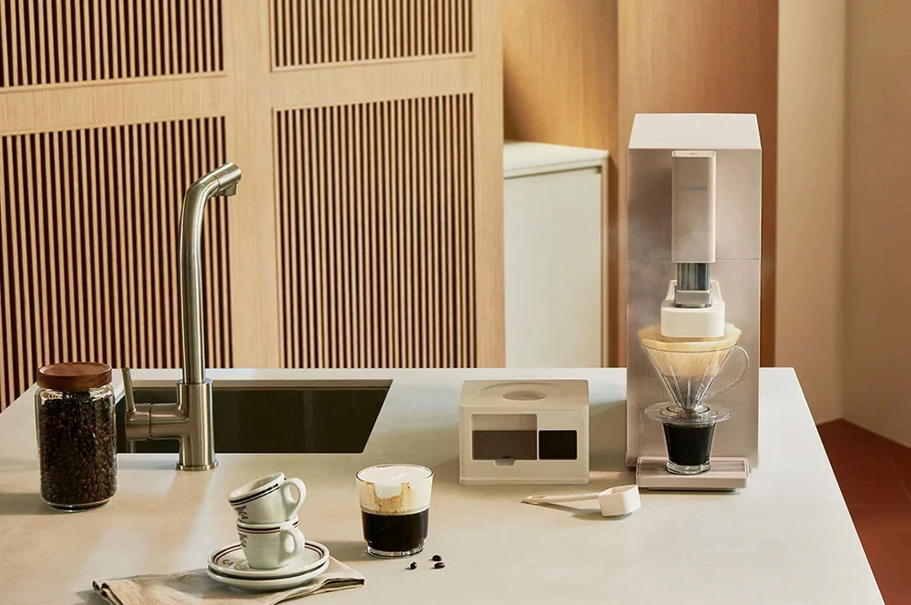 A STEAM 100 Barista water purifier setup on a light countertop in a minimalist kitchen-like interior. Visible items include a tall white and clear coffee maker with a glass cone filter, a small white box, a glass jar of coffee beans, two stacked ceramic cups, and a small glass cup with dark coffee.