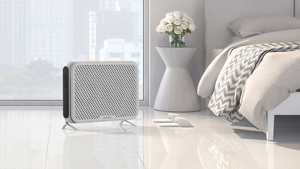 Ultra-Slim Wall-Mounted Air Purifier placed on the floor of a bright bedroom next to a bed and a small table with flowers. (CAC-AA0910FW)